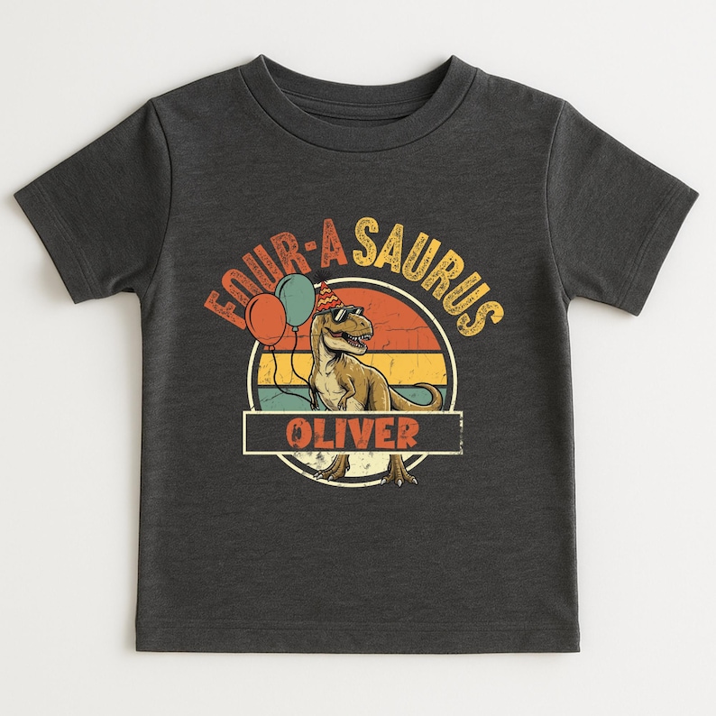 Detailed view of the nameplate customization on a dinosaur birthday shirt. The name "OLIVER" is written in a classic block font inside a vintage-style border. Above the name, the cool T-Rex with sunglasses signifies the "Four-a-Saurus" milestone. The distressed finish on the graphic provides a trendy, lived-in look for modern kids.