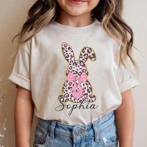 May include: A beige t-shirt featuring a pink and black leopard print bunny design with a pink bow and a glitter heart. The name "Sophia" is written in cursive below the bunny.