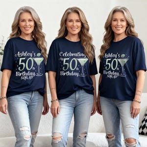 May include: Three women wearing navy blue t-shirts with a birthday design. The shirts feature a martini glass graphic and the text "Ashley's 50th Birthday Squad" or "Celebrating My 50th Birthday". They are wearing blue jeans.