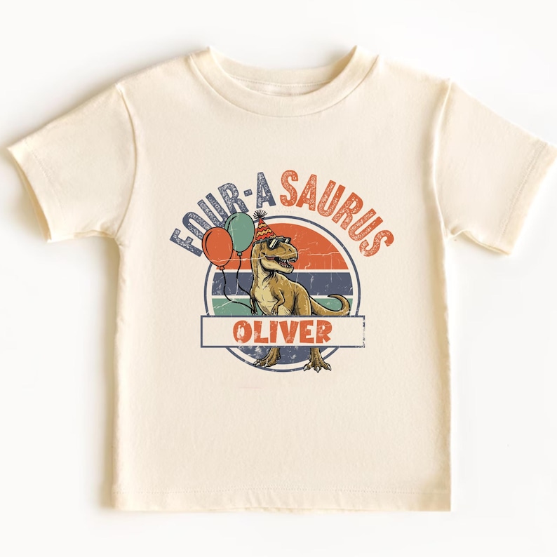 Detailed close-up of a personalized dinosaur birthday design on a soft toddler t-shirt. The text "FOUR-A-SAURUS" is arched in a vintage blue and orange font. The central T-Rex graphic has a textured green finish and is wearing "cool" black sunglasses. The name "OLIVER" is printed in a bold orange font inside a nameplate, making it a unique 4th birthday keepsake for boys.