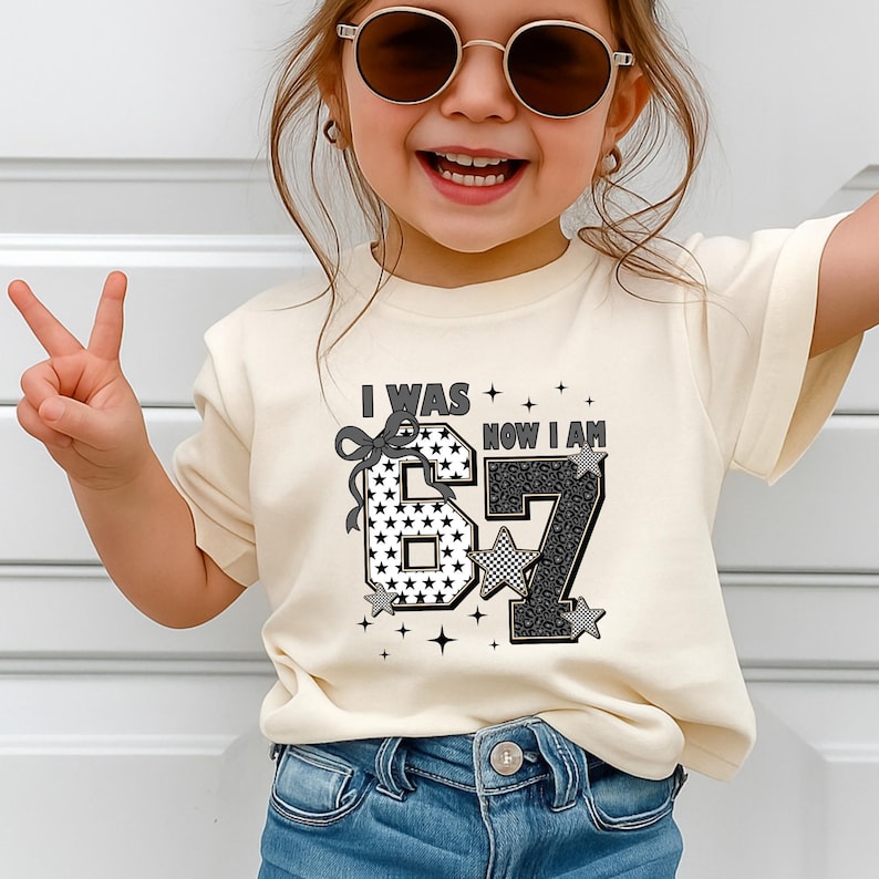 A lifestyle shot of a smiling girl modeling a 7th birthday outfit. She is wearing a pastel pink crewneck tee with the "I Was 6 Now I Am 7" graphic. The design highlights the transition from being six to turning seven. The aesthetic is preppy and fun, suitable for school parties or family gatherings. The shirt is paired with denim jeans, creating a casual yet stylish look for a young fashionista.