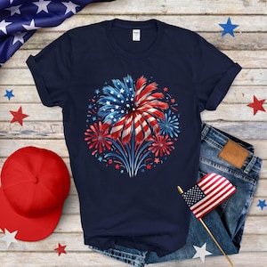 Op de afbeelding: Marineblauw T-shirt met een vuurwerkontwerp in rood, wit en blauw, dat doet denken aan de Amerikaanse vlag. Het ontwerp bevat ster- en streepuitbarstingen, ideaal voor patriottische evenementen. Het shirt wordt getoond met een spijkerbroek en een rode baseballpet.