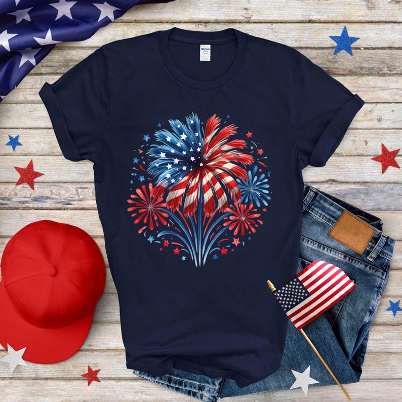 Op de afbeelding: Marineblauw T-shirt met een vuurwerkontwerp in rood, wit en blauw, dat doet denken aan de Amerikaanse vlag. Het ontwerp bevat ster- en streepuitbarstingen, ideaal voor patriottische evenementen. Het shirt wordt getoond met een spijkerbroek en een rode baseballpet.