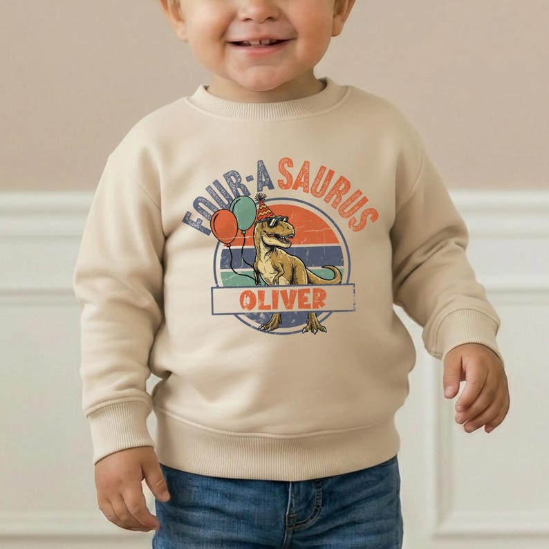 A happy toddler boy with a bright smile wearing a cream-colored sweatshirt with a "Four-A-Saurus" design. The sweatshirt features a green T-Rex wearing orange sunglasses and a party hat, standing in front of a retro yellow and orange sunset. The name "OLIVER" is personalized at the bottom. The child is posing with his hands in his pockets, highlighting a trendy 4th birthday milestone look.
