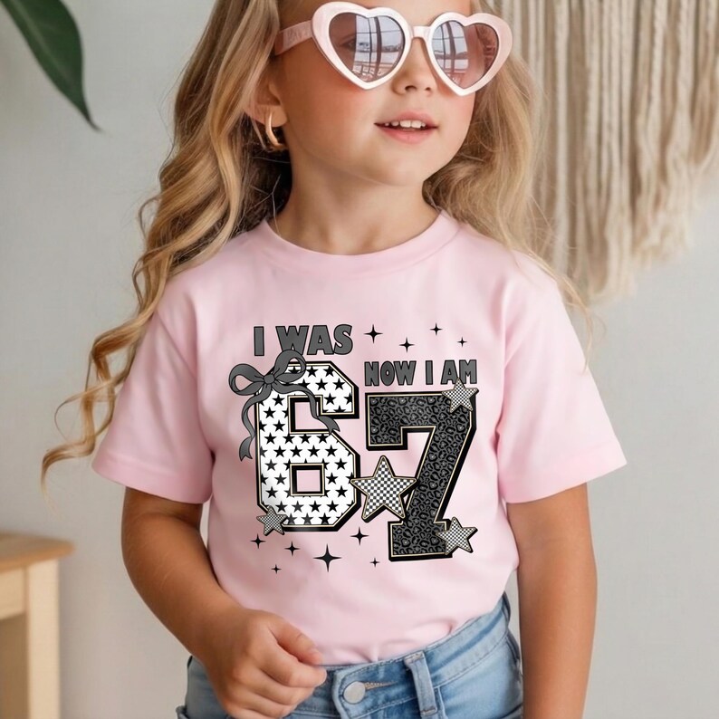 A cute young girl with blonde wavy hair wearing pink heart-shaped sunglasses and a light pink t-shirt. The shirt features a trendy birthday design that reads "I WAS 6 NOW I AM 7". The numbers are large; the number 6 has a white background with black stars, and the number 7 features a black and grey leopard print. A cute ribbon bow sits on the number 6. This outfit is perfect for a 7th birthday party or a second-grade celebration.