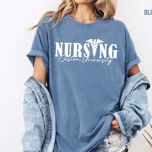 May include: Blue jean colored t-shirt with the word "NURSING" in white block letters and a medical symbol. Below the word "NURSING" is the text "Custom University" in a cursive font. The shirt is worn with a denim jacket.