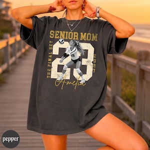 May include: A charcoal gray short-sleeve t-shirt featuring a graphic print with the text SENIOR MOM, THE FINAL SET, 23, 2027, and the name Amelia. The design includes a volleyball player in a white uniform and a volleyball in a distressed style.