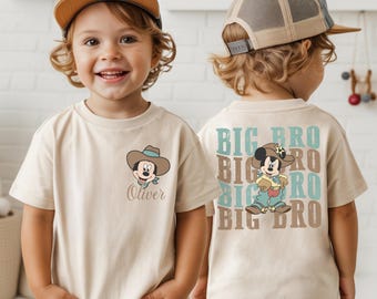 May include: Beige t-shirt featuring a cartoon Mickey Mouse wearing a cowboy hat and a blue bandana. The shirt has the name "Oliver" printed below the graphic. The back of the shirt says "BIG BRO" in a repeating pattern.