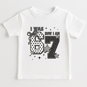 Close-up of a pink birthday shirt design for a seven-year-old girl. The text "I WAS" is above the number 6, and "NOW I AM" is above the number 7. The design uses mixed patterns like checkered squares, stars, and animal prints in black, white, and grey tones. Small star graphics surround the text. This graphic tee is a popular choice for milestone birthdays and trendy kids' fashion on Etsy.