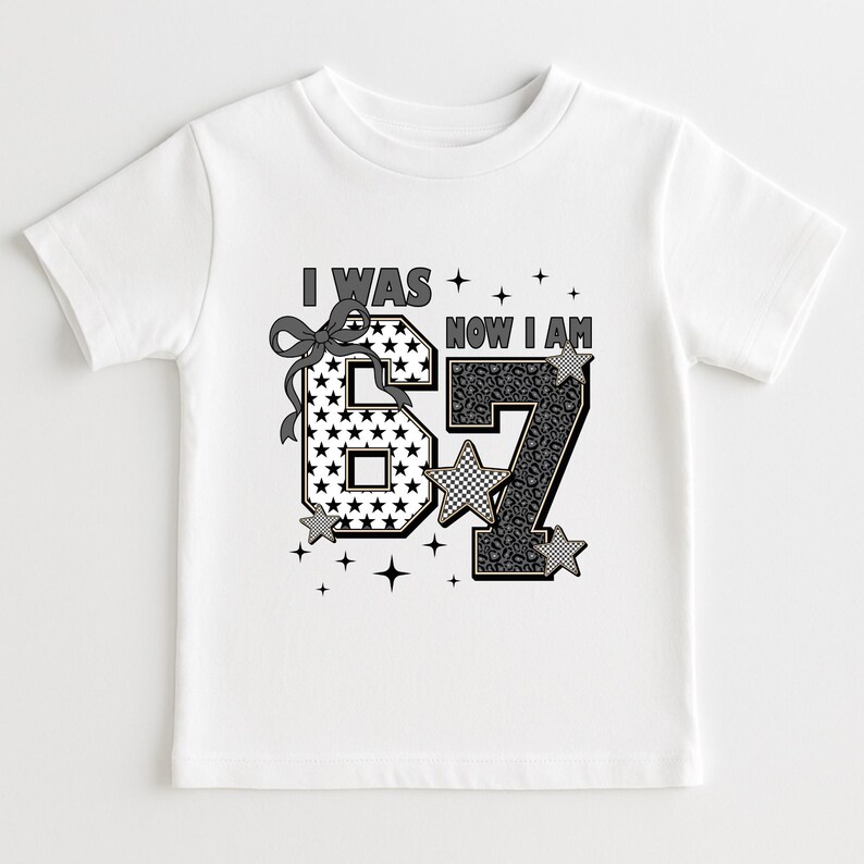 Close-up of a pink birthday shirt design for a seven-year-old girl. The text "I WAS" is above the number 6, and "NOW I AM" is above the number 7. The design uses mixed patterns like checkered squares, stars, and animal prints in black, white, and grey tones. Small star graphics surround the text. This graphic tee is a popular choice for milestone birthdays and trendy kids' fashion on Etsy.