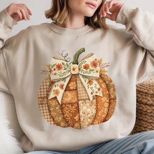 May include: Beige sweatshirt featuring a large, patchwork pumpkin design. The pumpkin is composed of various fabrics in shades of orange, brown, and cream, with a floral bow and green stem. A fall-themed apparel item.