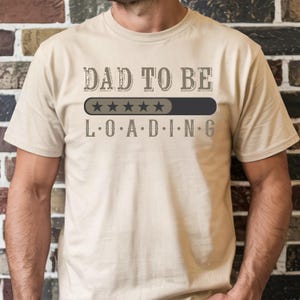 May include: Beige t-shirt with the text "DAD TO BE LOADING" in a distressed font. The design includes a loading bar with five stars, and the letters L.O.A.D.I.N.G. below. The shirt is short-sleeved.