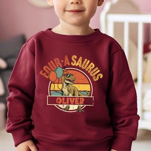Close-up on the typography and graphics of a personalized dinosaur hoodie for boys. The phrase "FOUR-A-SAURUS" is a fun play on words for a 4th birthday. The print is sharp and professional, showing the fine details of the T-Rex's scales and the reflections on its cool black sunglasses. Perfect for a dinosaur-loving four-year-old.