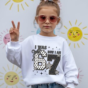 A young girl wearing a white sweatshirt with the number 67 and the text "I WAS 66 NOW I AM" is making a peace sign.