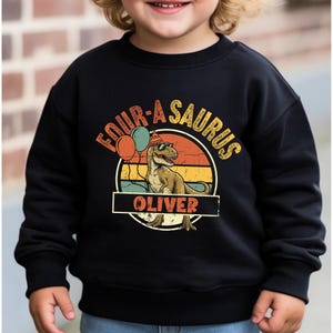 Full view of the "Four-A-Saurus" toddler tee showing the fit and high-quality fabric. The design includes a large dinosaur graphic with party balloons and a retro sunset. The alt text emphasizes the "Custom Name" and "4th Birthday" keywords, making it easy for Etsy shoppers to find this unique birthday outfit for their son.