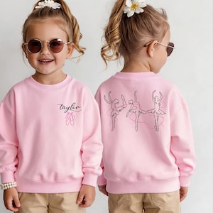 May include: Pink sweatshirt with the name "Taylor" in cursive and a pair of pink ballet shoes on the front. The back features a line drawing of three ballerinas. A cute and stylish design for young dancers.