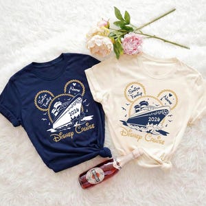 May include: Two navy blue and cream-colored t-shirts with a Disney Cruise design. The design features a cruise ship, Mickey Mouse ears, and the text "Disney Cruise 2024". A bottle of wine and flowers are in the image.