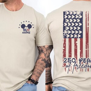 A beige t-shirt with a graphic design on the back, depicting an American flag and Mickey Mouse ears. The design also includes the text "250 YEARS OF FREEDOM" and the person's name "NAME".