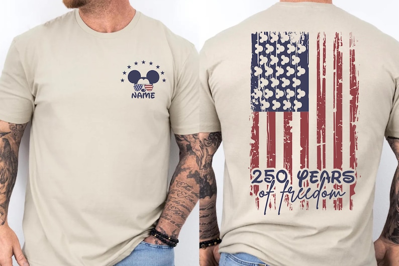 A beige t-shirt with a graphic design on the back, depicting an American flag and Mickey Mouse ears. The design also includes the text "250 YEARS OF FREEDOM" and the person's name "NAME".