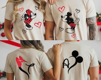 May include: Beige t-shirts featuring Mickey and Minnie Mouse designs. The front of the shirts display Mickey and Minnie Mouse with red hearts. The back of the shirts show a Minnie Mouse bow and a Mickey Mouse silhouette with the letter "A".