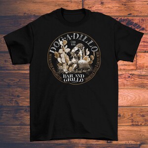 May include: Black t-shirt with a circular graphic featuring the text "Dogadillo Bar and Grillo" and illustrations of a desert scene with cacti and a tortoise. The shirt also includes the text "Ciudad Acuna" and "Prickly Pear Margaritas".