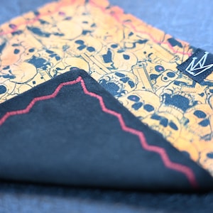 May include: A black and orange pocket square with a skull pattern and red stitching. The pocket square has a small black label with a crown logo.