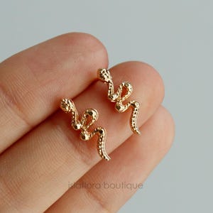 May include: A pair of gold-toned earrings with a delicate, twisted design. The earrings are made of small, round beads that are connected together to form a curved shape.