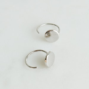 May include: A pair of silver hoop earrings with a circular disc pendant. The earrings are simple and elegant.