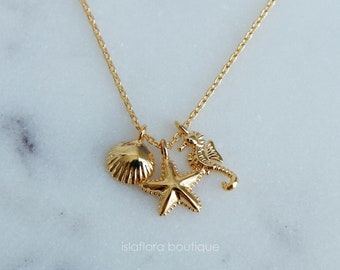 May include: Gold necklace with a seashell, starfish, and seahorse charm. The necklace is on a white marble background.