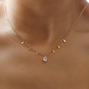 May include: Gold-toned necklace with a delicate chain and multiple small leaf-shaped charms. A teardrop-shaped, clear gemstone pendant hangs at the center. The necklace is worn against a light-colored top.