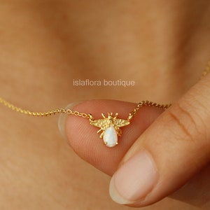 May include: A delicate gold necklace featuring a bee pendant with a white opal-like stone body. The bee has intricate wing and body details. The necklace is held by a person's hand, showcasing the fine chain and pendant design.