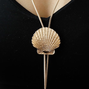 May include: Gold-toned seashell pendant necklace. The pendant is a detailed, textured seashell design. The necklace has a long, delicate chain that hangs down. The background is a solid black.