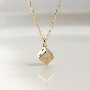 May include: A gold-colored necklace with a small, three-dimensional die pendant. The die has visible dot markings on its sides. The chain and pendant are reflective against a white background. The necklace is a jewelry item.