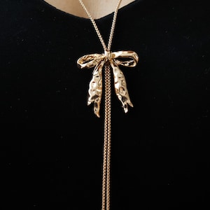May include: Gold-toned necklace featuring a textured bow pendant. The bow has a hammered metal appearance and is suspended from a delicate chain. The necklace has two long, dangling chains. The background is a solid black.