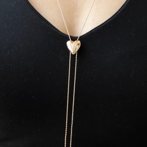 May include: A gold-colored heart-shaped pendant necklace with a delicate chain. The necklace features a larger heart pendant at the neckline and two dangling chains with small heart charms. The necklace is displayed against a black background.