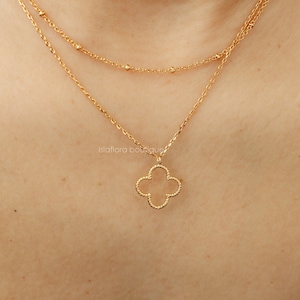 Könnte beinhalten: Eine goldene Halskette mit einem zarten vierblättrigen Kleeblatt-Anhänger. Die Kette ist mit zwei Ketten geschichtet, eine mit kleinen Perlen und die andere mit einer dickeren Kette.