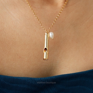 May include: A gold necklace with a whistle pendant and a single pearl charm. The necklace is worn by a person with a blue shirt.