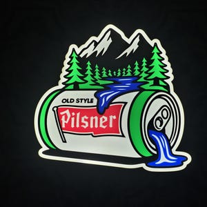 Peut inclure: Autocollant représentant une canette de bière Pilsner avec un paysage de montagne et de forêt. La canette est blanche avec des accents verts et le texte "Old Style Pilsner". Du liquide bleu coule de la canette.