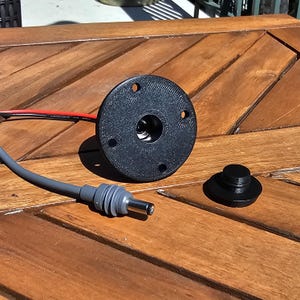 May include: A black electrical outlet with a round, flat faceplate and four screw holes. A grey electrical cord with a plug is connected to the outlet. A separate black round electrical cover is also shown.
