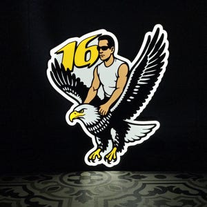 May include: A sticker showing a man in sunglasses riding a bald eagle. The eagle is black and white with yellow details. The number "16" is in yellow. The background is black.