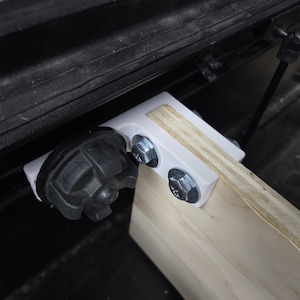 May include: A white 3D-printed bracket secures a light-colored wooden board to a black metal frame. The bracket features a circular black knob on one side and is fastened to the wood with two silver-colored hexagonal bolts and matching nuts.
