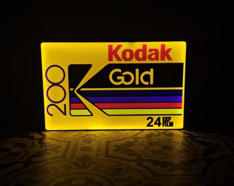 May include: A glowing yellow sign featuring the Kodak Gold logo in red and gold. The sign also displays the numbers "200" and "24 EXP" in white. The design includes a black rectangle with colorful stripes.