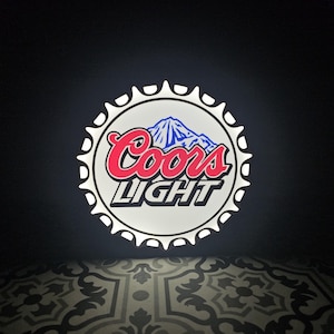 Peut inclure: Un panneau circulaire en forme de capsule de bouteille, avec le logo Coors Light. Le logo comprend le nom de la marque en rouge, un graphique de montagne et le mot "LIGHT" en blanc. Le panneau est éclairé sur fond sombre.