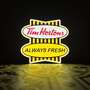 May include: A Tim Hortons sign with a white border and a yellow and brown striped design. The top oval features the words "Tim Hortons" in red script. The bottom oval reads "ALWAYS FRESH" in white.