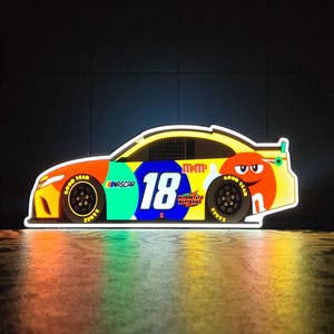 Peut inclure: Une enseigne murale LED lumineuse en forme de voiture de course NASCAR. La voiture présente un design jaune, orange et bleu avec le numéro 18, la marque M&M's, le logo Interstate Batteries et le texte NASCAR. L'enseigne émet une lumière vive.