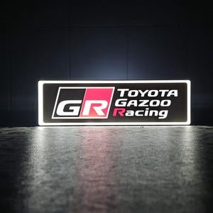 Puede incluir: Letrero rectangular iluminado con el logotipo de Toyota Gazoo Racing. El letrero tiene un borde negro, letras blancas y un logotipo GR rojo y negro. El texto "TOYOTA Gazoo Racing" se muestra en blanco.