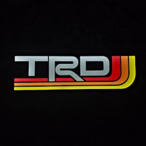 Puede incluir: El logotipo de TRD, en estilo retro, se muestra sobre un fondo negro. Las letras TRD son plateadas, con un diseño de rayas rojas, naranjas y amarillas. El logotipo es un elemento de diseño.