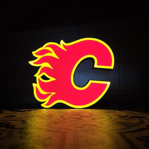 May include: A neon sign in the shape of the letter "C" with a flame design, in red and yellow. The sign is illuminated against a dark background, with a patterned surface below.