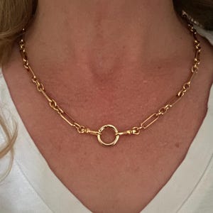 May include: A gold-colored necklace with a large circular clasp and a chain of alternating rectangular and oval links. The necklace is worn around the neck and rests on a white v-neck shirt.