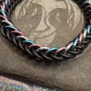 May include: A black, blue, pink, and silver chainmail bracelet. The bracelet is made of metal rings that are woven together in a pattern. The bracelet is lying on a gray stone surface.